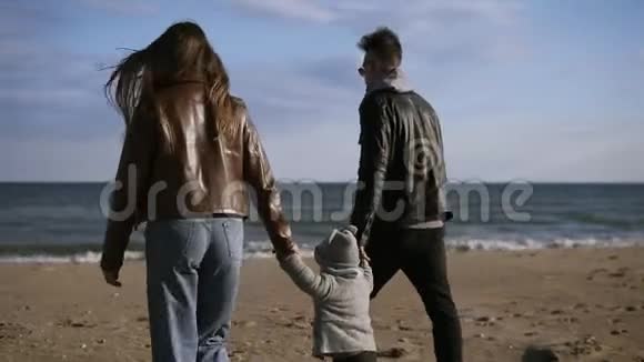 时髦年轻的父母带着儿子走在中间正走在秋天风起的海边手握双手视频的预览图