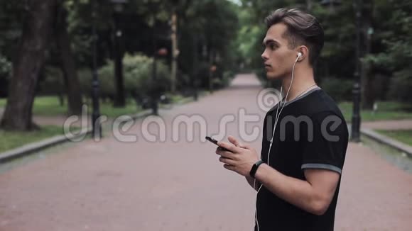 有魅力的年轻人戴着耳机手里拿着智能手机站在公园里转动他的头部理念健康视频的预览图