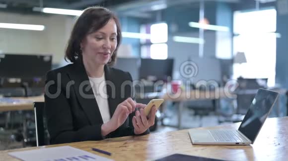 成功的中年女商人使用智能手机庆祝视频的预览图
