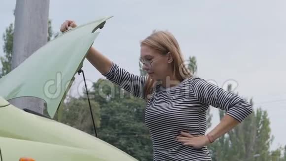 一个年轻的女人站在一辆开着引擎盖的车旁边看着引擎汽车在行驶中突然抛锚了视频的预览图