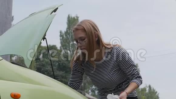 一个年轻的女人站在一辆开着引擎盖的汽车旁边用油尺检查发动机的油位视频的预览图