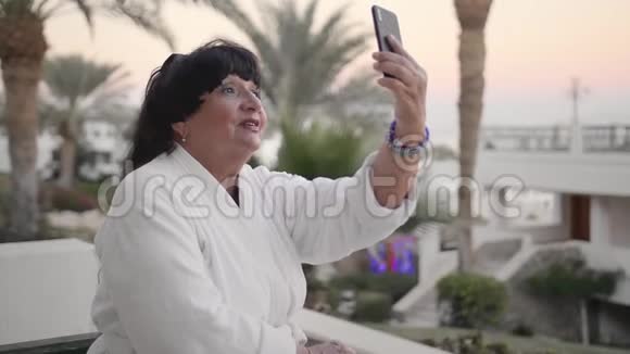 穿着白色特里长袍的快乐白种人高级女性正在一个热带度假胜地休息并打了一个视频电话使用视频的预览图