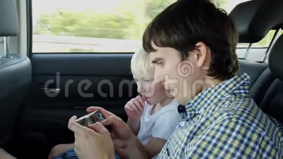 父亲和儿子坐在汽车座椅上一边玩智能手机一边在后座上开车视频的预览图
