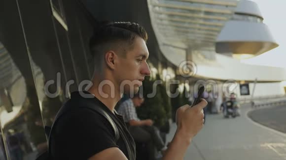 迷人的年轻人穿着黑色t恤使用他的智能手机录音和滚动在机场或汽车站视频的预览图