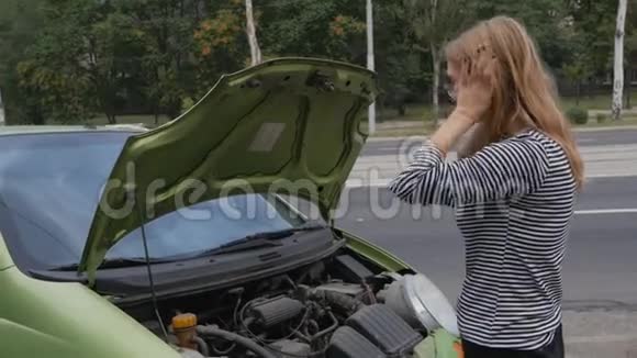 一个年轻的女人站在一辆开着引擎盖的车旁边看着引擎汽车在行驶中突然抛锚了视频的预览图