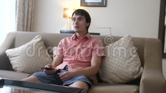 年轻的帅哥在看电视男人坐在沙发上惊讶地张开嘴捂着脸视频的预览图