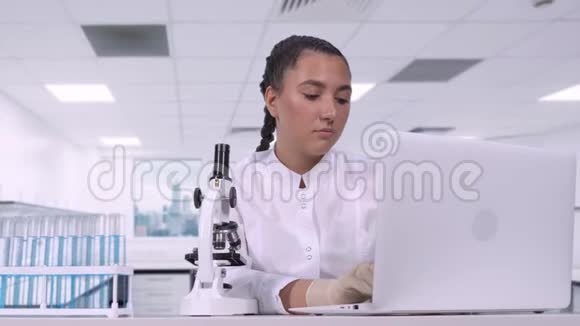 一位女性研究员在显微镜下观察生物样本并将研究结果写在笔记本电脑上视频的预览图