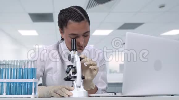 一位女性研究员在显微镜下观察生物样本进行临床试验并将数据写到笔记本电脑上视频的预览图