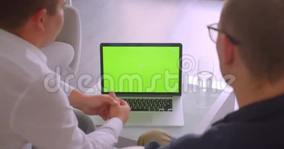 两位商人使用带有绿色屏幕的笔记本电脑坐在扶手椅上举行会议视频的预览图