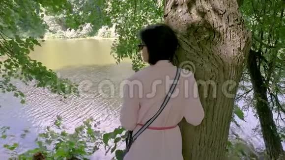 一位中年妇女靠在一棵树旁的池塘边休息视频的预览图