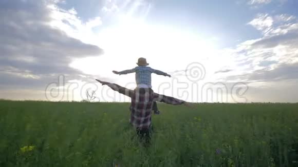 幸福的家庭儿子坐在父亲的肩膀上玩着举起的手臂走在背景天空的油菜田上视频的预览图