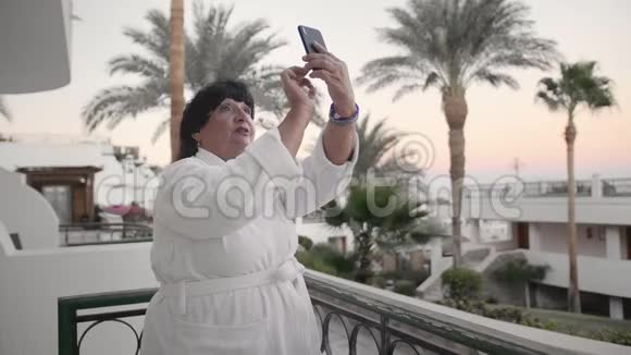 白人高级女性在智能手机上拍照它应该在酒店的露台上穿着白色的特里长袍视频的预览图