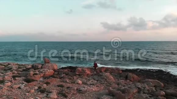 年轻的女摄影师正在寻找一个地方在美丽的岩石海岸拍摄照片日落时有巨大的海浪幼仔视频的预览图