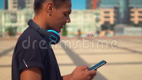 侧视时髦的家伙带着耳机手持智能手机在城里散步视频的预览图