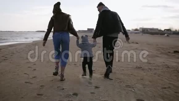 一个年轻的家庭在一个刮风的日子沿着海边散步丈夫和妻子牵着一个小儿子的手视频的预览图