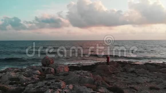 迷人的白种人女孩站在岩石海岸的大石头上在日落时拍摄美丽的地中海医生视频的预览图