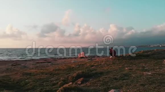 年轻迷人的女摄影师坐在岩石海滩上拍摄粉红色美丽的日落和壮丽的多云天空视频的预览图