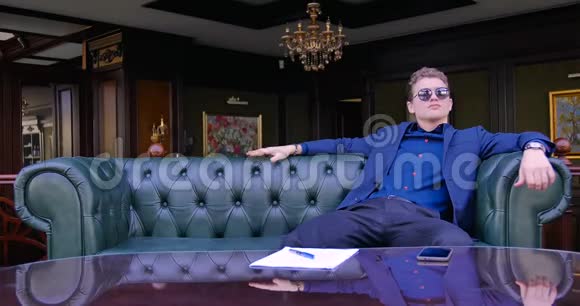 一个年轻的商人穿着蓝色的西装和眼镜坐在豪华公寓的皮椅上等待视频的预览图