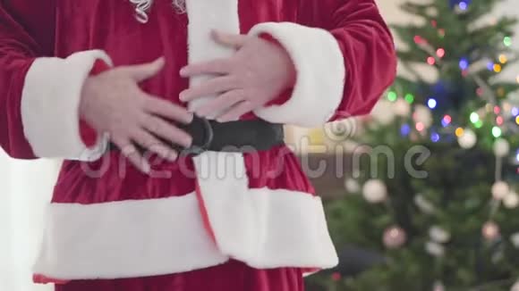 穿着圣诞老人服装的不可辨认的白人男人抚摸着他的肚子在圣诞树前跳舞概念视频的预览图
