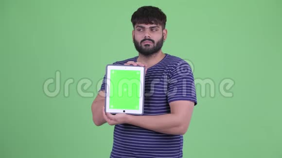 一位年轻体重超重留着胡子的印度男人一边看着数码平板电脑一边思考视频的预览图