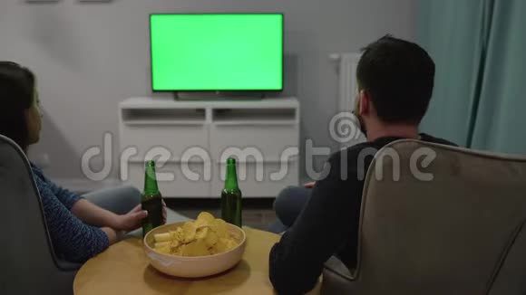 男人和女人坐在椅子上用绿色的屏幕看电视喝啤酒吃薯片讨论他们看到了什么视频的预览图