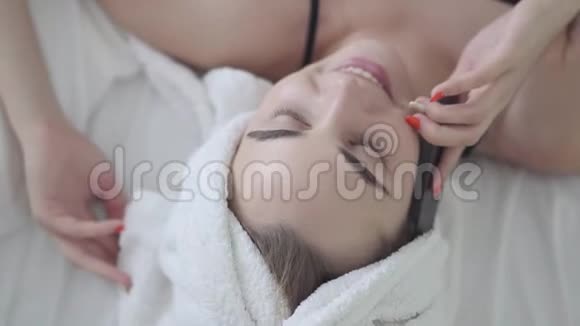 可爱的年轻女子头上拿着毛巾躺在床上说话手机在家休闲休息日视频的预览图