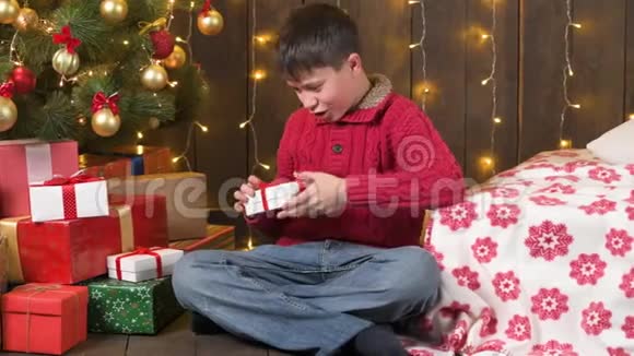 男孩坐在室内靠近装饰的圣诞树灯光和礼物圣诞快乐和节日快乐视频的预览图