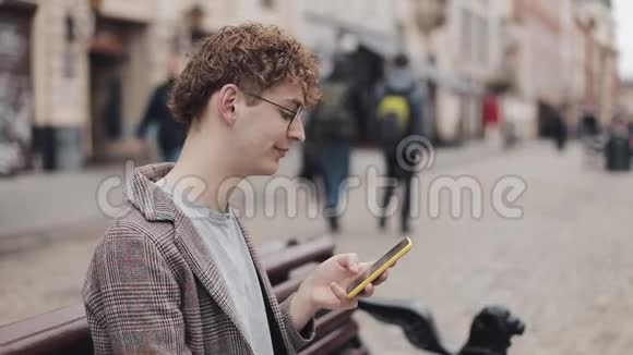 戴眼镜和夹克的年轻嬉皮士的侧视使用他的智能手机在屏幕上滚动一边微笑视频的预览图