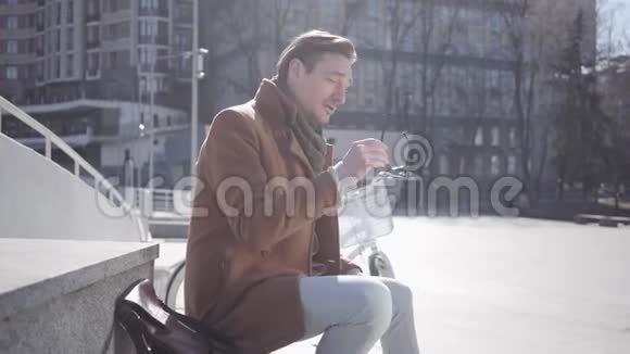 一个戴着太阳镜的时尚男子坐在城市建筑背景下的肖像视频的预览图