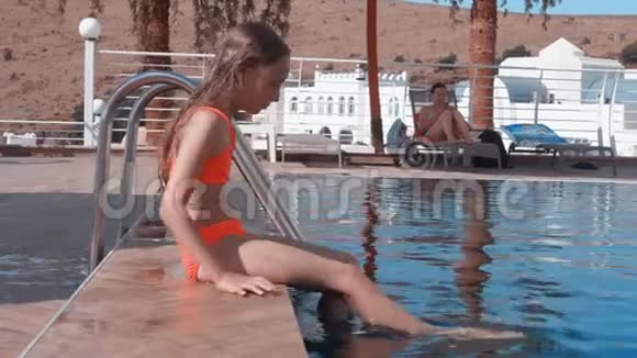 在度假酒店里坐在池边的少女用双腿蘸水小女孩在游泳池旁泼水视频的预览图