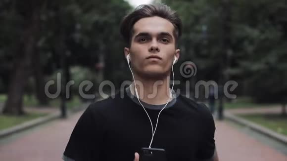 帅气的年轻人戴着耳机手拿着智能手机在公园里奔跑的肖像亲密的概念健康视频的预览图