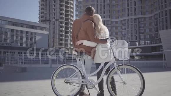 高个子男人抱着漂亮的金发女人穿着温暖的夹克站在自行车旁边看着高楼这对夫妇在聊天视频的预览图