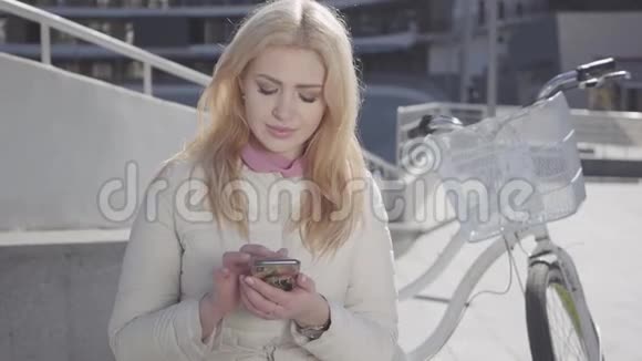 漂亮的金发女人穿着温暖的白色夹克坐在城市街道上骑着自行车在手机上打字妇女视频的预览图