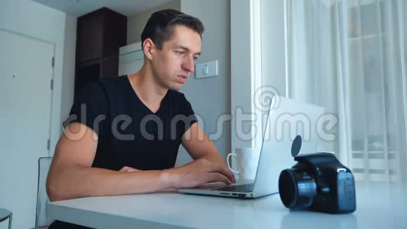 年轻的摄影师使用笔记本电脑处理图像在他的笔记本电脑和喝咖啡在家里外部硬盘摄像头视频的预览图