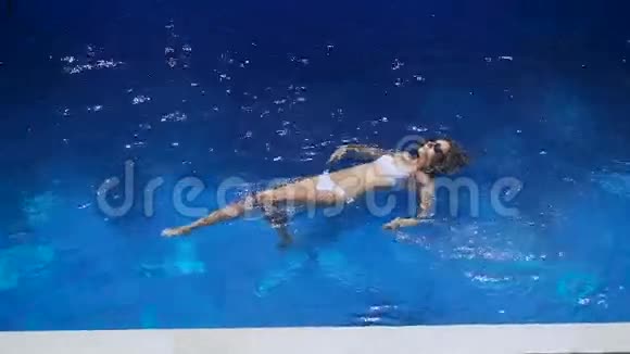 一个穿着白色泳衣的瘦女孩在游泳池里背上漂浮美丽的年轻女子在度假视频的预览图