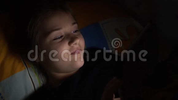 可爱的小女孩躺在床上看着一个没有周围灯光的平板电脑或电话只看到她脸上的倒影视频的预览图