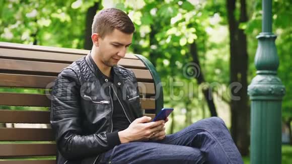 有魅力的家伙在公园里坐在长凳上用智能手机视频的预览图