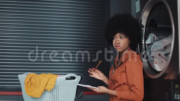 年轻有魅力的非洲裔美国妇女写做清单在记事本上坐在自助公共洗衣视频的预览图