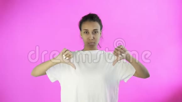 穿着白色T恤的漂亮少女拇指朝下双手放在粉色背景上视频的预览图