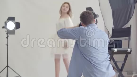 拍摄的后台摄影师在摄影棚里拍摄站在白色背景上的迷人女人的照片视频的预览图