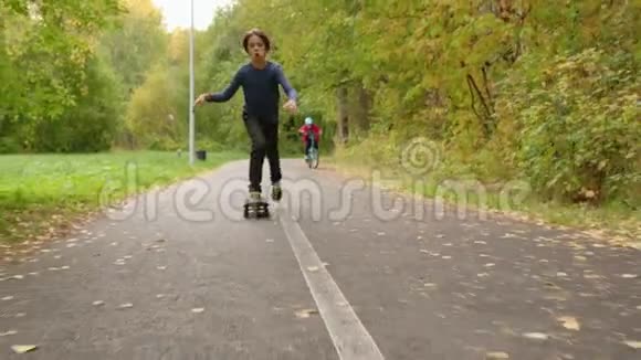 在公园路上骑滑板的少年男孩在秋天公园骑自行车的小男孩滑板和自行车视频的预览图