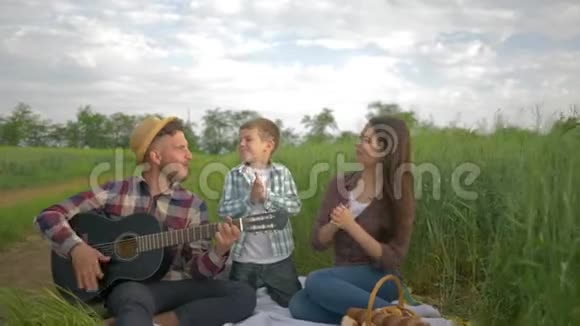 快乐的家庭快乐的爸爸弹吉他妈妈带着儿子唱歌拍手在绿色的田野里放松地野餐视频的预览图