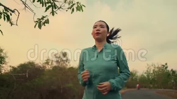 慢速跟踪万向节拍摄年轻健康的亚裔韩国妇女在日落时分在树林中跑步做慢跑锻炼视频的预览图
