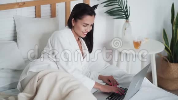 穿着浴袍的年轻女子躺在床上用笔记本电脑在网上工作或学习视频的预览图