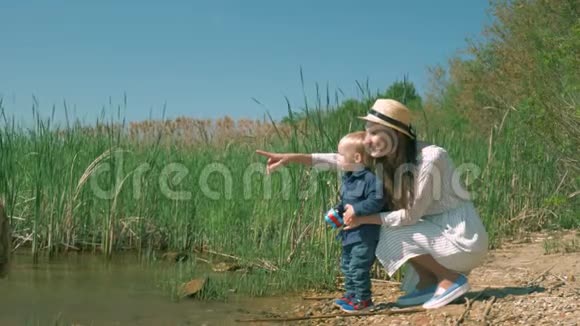 年轻快乐的妈妈在用芦苇静静地躺在岸边的湖上向小男孩展示大自然视频的预览图