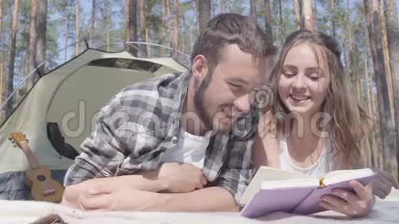 肖像胡子男和可爱的年轻女子躺在森林里的帐篷里看书爱视频的预览图