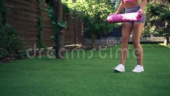 年轻女子在绿草上卷起粉色健身垫慢动作视频的预览图