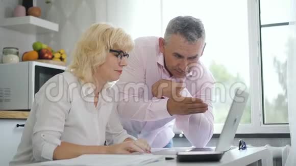 男性养老金领取者教退休妻子在现代厨房使用笔记本电脑视频的预览图