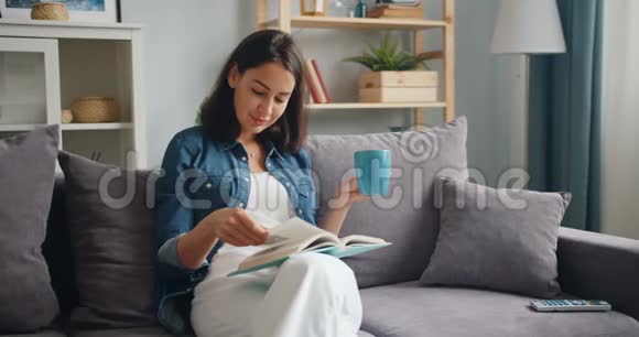 聪明的年轻女士拿着一杯茶坐在公寓的沙发上看书视频的预览图