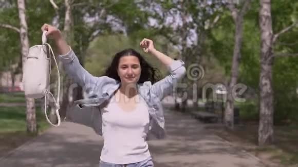 风和日丽的天气里一个手拿背包留着长发的滑稽女孩走在街上跳舞很滑稽视频的预览图